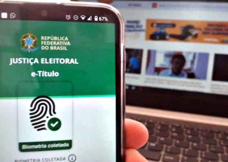 Imagem mostra celular com aplicativo do título de eleitor e ao fundo um computador