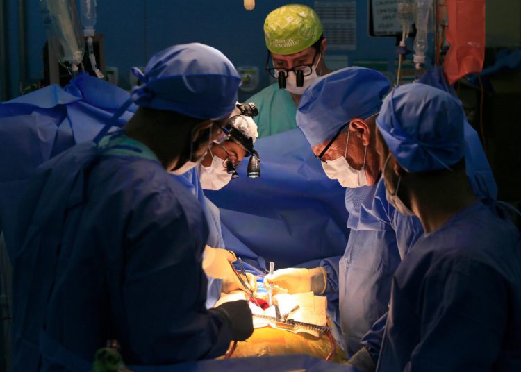 sala de cirurgias com procedimento em andamento, vários médicos rodeiam ponto luminoso portando bisturis