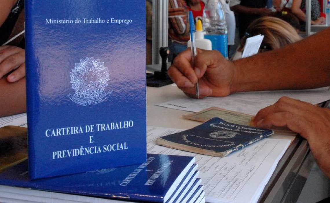 pilha de carteiras de trabalho em foco. Ao fundo, homem assina papel com carteira de trabalho em mãos