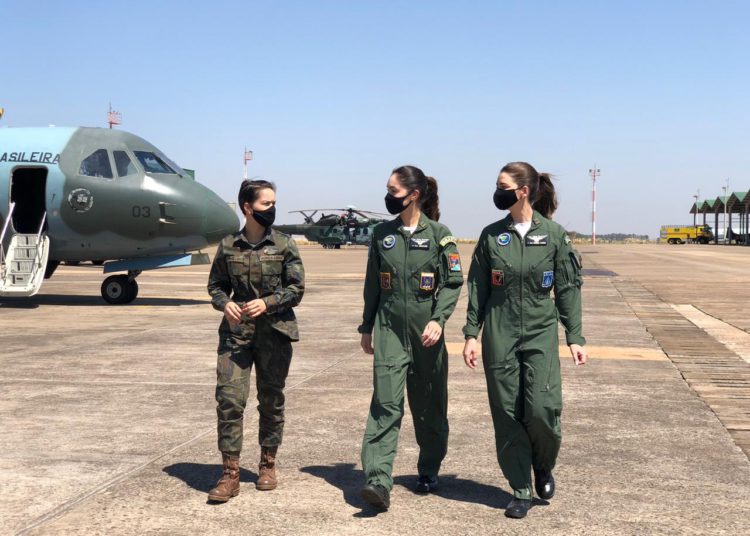 três integrantes mulheres da Força Aérea Brasileira conversando, com um avião ao fundo