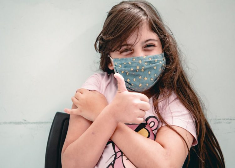 vacinada e com mão no braço, criança faz sinal de jóia com a mão