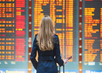 Imagem tem uma mulher segurando mala e diante de um painel de aeroporto