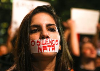 foto de mulher, durante protesto contra o feminicídio e as demais violências contra a mulher, com uma faixa tampando a boca, com os dizeres: o silêncio mata