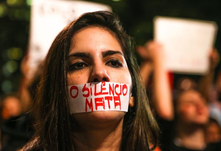 foto de mulher, durante protesto contra o feminicídio e as demais violências contra a mulher, com uma faixa tampando a boca, com os dizeres: o silêncio mata