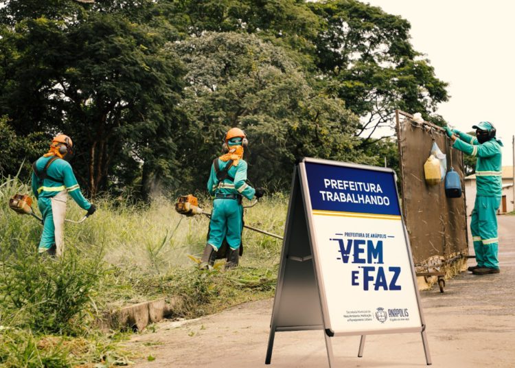 servidores municipais roçando terreno, com placa a frente escrito "projeto vem e faz"