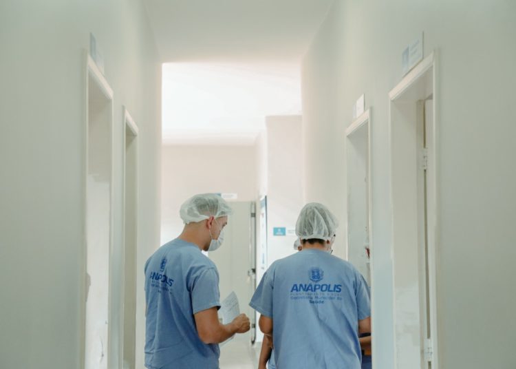 foto de dois profissionais da saúde, vestindo jaleco azul, de costas em corredor branco