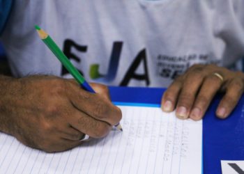 foto de pessoa, com a camisa da EJA (Educação para Jovens e Adultos), escrevendo em caderno
