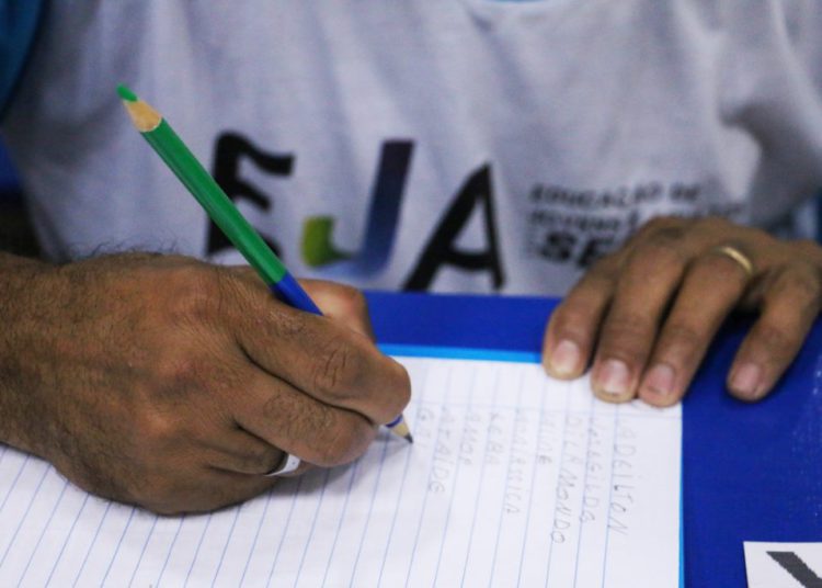 foto de pessoa, com a camisa da EJA (Educação para Jovens e Adultos), escrevendo em caderno