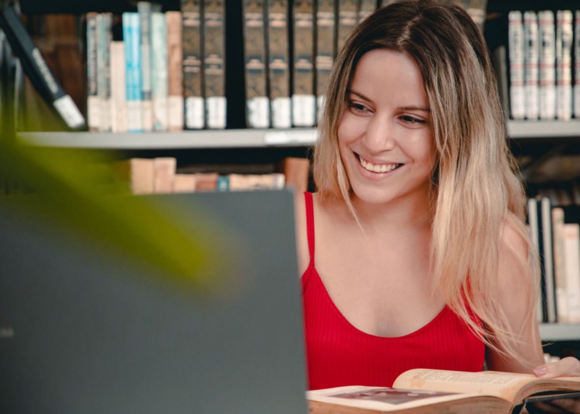 foto de estudante, sorrindo, olhando para o notebook e com livro aberto em mãos