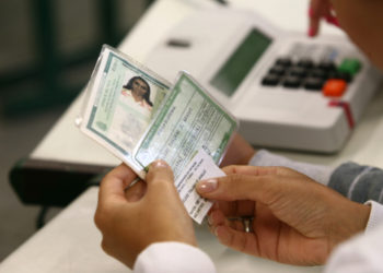 foto de título de eleitor nas mãos de mesária, ao fundo uma urna eleitoral