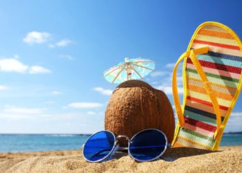 foto de óculos, coco e chinelo cravados em areia de praia, representando férias