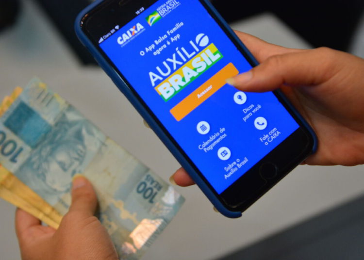 foto de celular aberto no aplicativo do auxílio brasil, com dinheiro em mãos ao lado