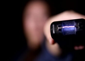 An out of focus woman on dark black background holds a stun gun up close to camera and fires it.