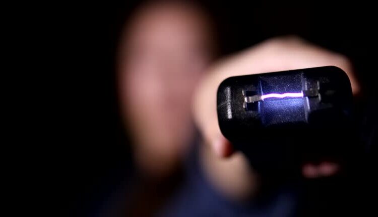 An out of focus woman on dark black background holds a stun gun up close to camera and fires it.