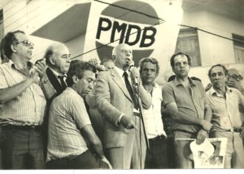 O deputado federal Ulysses Guimarães discursa, tendo ao seu lado à direita: Iris Rezende, Adhemar Santillo e Anapolino de Faria.
No canto esquerdo, o ouvinte é Henrique Santillo.