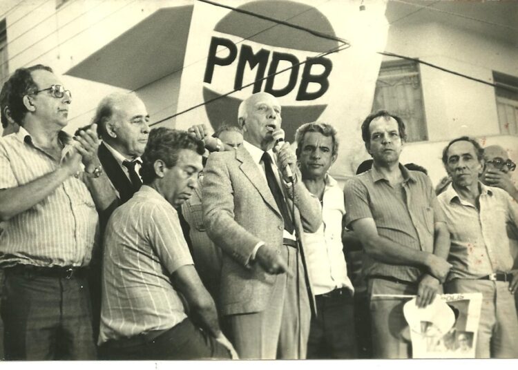 O deputado federal Ulysses Guimarães discursa, tendo ao seu lado à direita: Iris Rezende, Adhemar Santillo e Anapolino de Faria.
No canto esquerdo, o ouvinte é Henrique Santillo.