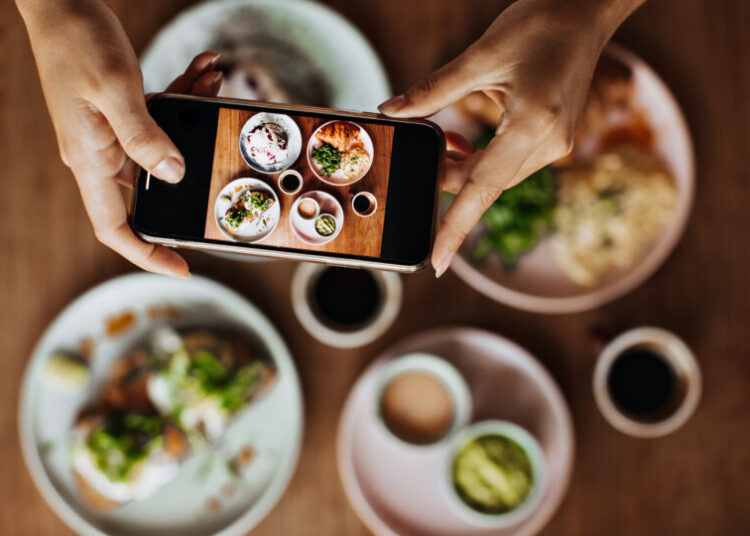 Snapshot of tanned female hands holding smartphone and taking photos of plate with meal. Picture of delicious colorful food on wooden table